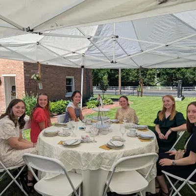 Youth sitting at a tea party table with green grass and orange brick whitaker home in the background on a sunny summer day